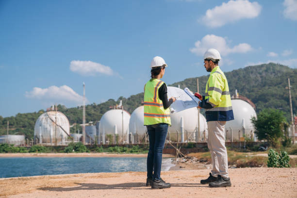 istock Two engineers in safety gear with blueprints discussing work in front of industrial storage tanks on a clear day. 1995113497