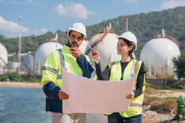 istock Two engineers in safety gear with blueprints discussing work in front of industrial storage tanks on a clear day. 1995116275