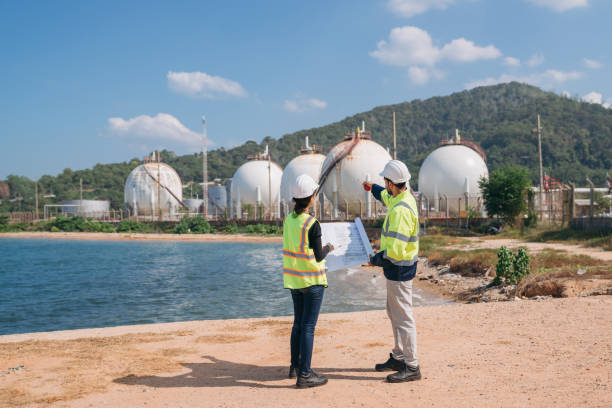 istock Two engineers in safety gear with blueprints discussing work in front of industrial storage tanks on a clear day. 1995117977
