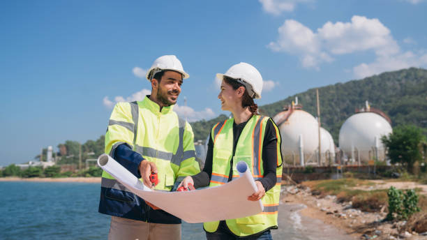 istock Two engineers in safety gear with blueprints discussing work in front of industrial storage tanks on a clear day. 1995118564