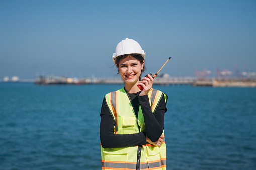 A young female engineer wearing a safety helmet and reflective vest holds a walkie-talkie at an industrial storage facility.