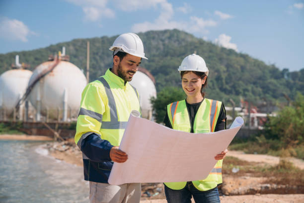 istock Two engineers in safety gear with blueprints discussing work in front of industrial storage tanks on a clear day. 1995123814