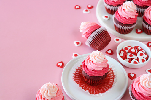 Stock photo showing close-up view of a batch of freshly baked, homemade, red velvet cupcakes in paper cake cases on and surrounding a cake stand. The cup cakes have been decorated with swirls of ombre effect pink piped icing and sugar hearts.  Valentine's Day and romance concept.