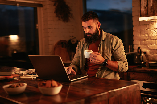 A young man is at home having a cup of coffee and working on his laptop late in the evening.