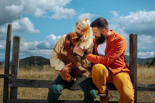A young Caucasian couple is on a weekend trip in the mountains enjoying their time with their dog on a meadow and sitting on a fence.