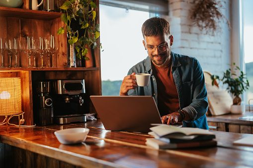 A young Caucasian businessman is working from home and having a cup of coffee as he's sitting at his computer.