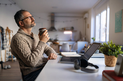Mature Caucasian man enjoying first morning coffee while working from his home office.