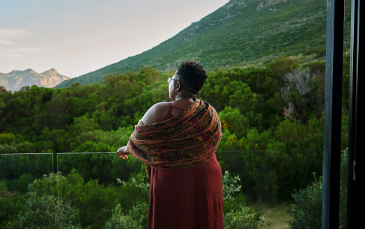 Rear view of a woman standing on the balcony of a hotel and spa and enjoying the scenic landscape view at sunset