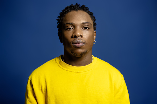 Portrait of thoughtful young african man staring at camera. African male in yellow t-shirt staring at camera against blue background.