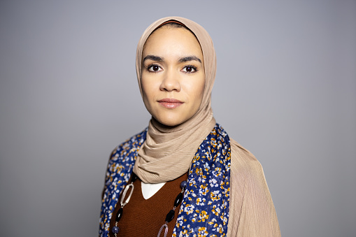 Portrait of young Islamic woman with blank expression on gray background. Muslim female with hijab staring at camera in studio.