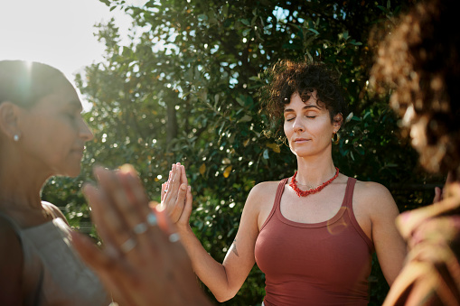 Young woman and a group of other women standing with their eyes closed with hands together during an exercise at a wellness retreat in summer