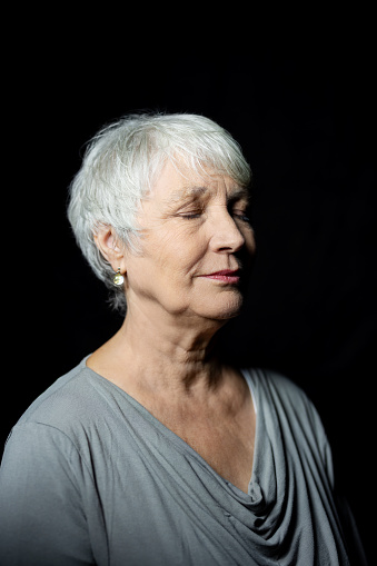 Portrait of senior woman with eyes closed. Mature female with short grey hair thinking against black background.