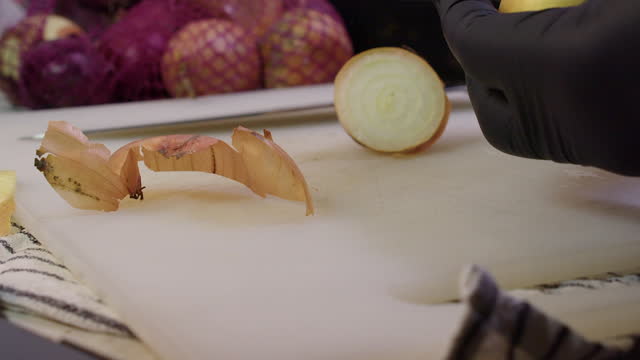 Gloved hands of chef cut and peel outer skin off onion bulb in kitchen