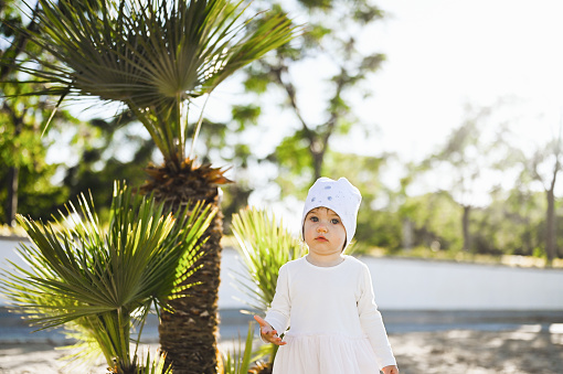 Toddler girl standing on a sandy beach near a palm tree.