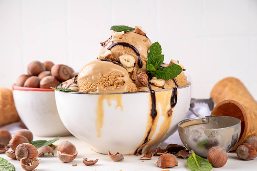 Hazelnut chocolate ice cream. Big bowl portion with nuts gelato scoops, chocolate topping and a lot of hazelnuts on kitchen table background