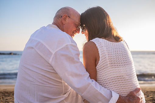 Rear view of bonding senior couple hugging while sitting on the beach face the sunset enjoying vacation and retirement, two elderly people expressing love and tenderness