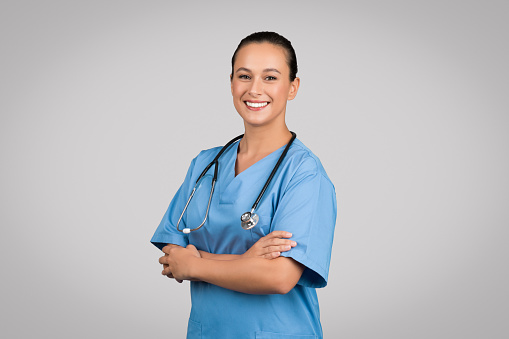 Happy nurse lady with folded arms posing wearing blue workwear and standing on gray studio background, smiling to camera. Healthcare and medicine concept