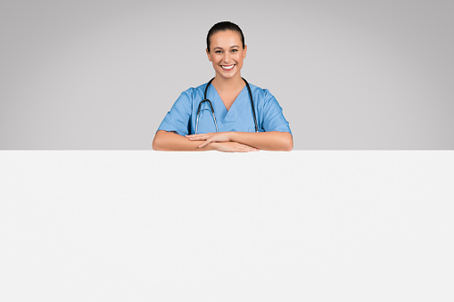 Medical ad. Cheerful european female doctor leaning on big blank advertising board with free space for your text or offer, posing over grey background, banner
