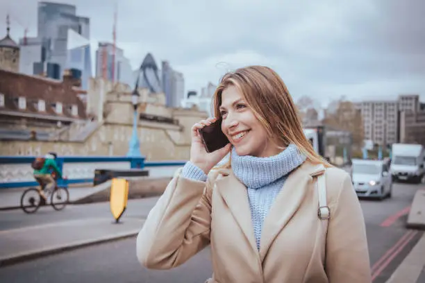 Candid photo of a young cheerful woman talking on her mobile phone in down town London, England. The girl is blond with warm clothes not looking into the camera and image is created with selective focus with sharpness priority over the model - creative stock photo.
