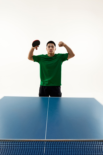 Exultant Asian man, table tennis champion demonstrated muscles in the end of match against white studio background. Young league. Concept of sport, hobby, lifestyle, match, victory, championship.
