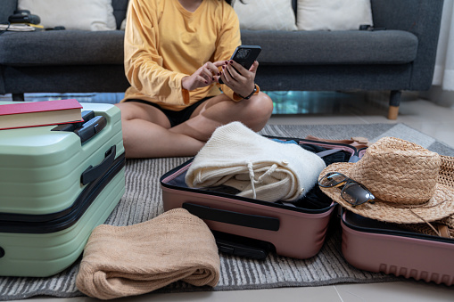 Young Asian Woman packing her suitcase for summer travel and checking hotel reserve on mobile phone