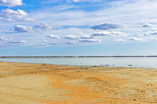 Beautiful shore of lake, white clouds on sky, water surface and sandy beach. Tranquil nature scene, summer sunlight day. Vibrant colored landscape in minimal style, panoramic cloudscape with skyline