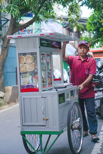 Jakarta, Indonesia - April 19, 2023: A burger vendor walks along the street peddling his wares. Indonesian street food. Small business entrepreneur.