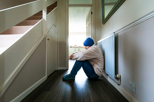 Color image depicting a mid adult man sitting on the floor next to a radiator at home. He is cold and is dressed in a beige fleece and knit hat. The man appears depressed and lonely with his knees drawn up into his body and his head bowed.
