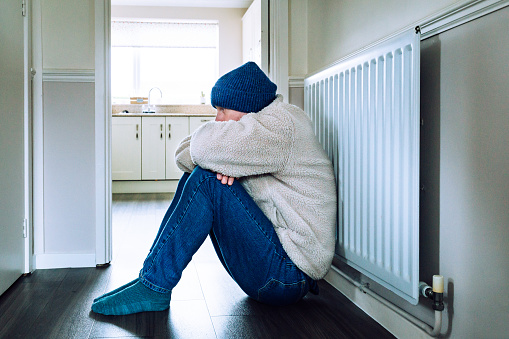 Color image depicting a mid adult man sitting on the floor next to a radiator at home. He is cold and is dressed in a beige fleece and knit hat. The man appears depressed and lonely with his knees drawn up into his body and his head bowed.