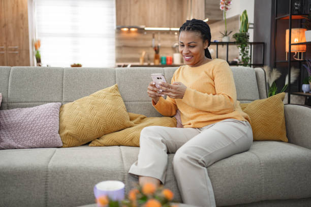 istock Happy black woman using mobile phone while sitting on a sofa at home 1995424797