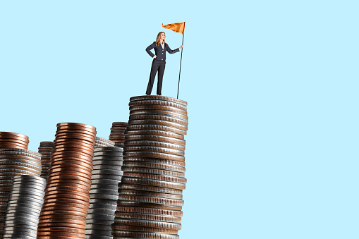A woman stands while holding a flag on top of a tall stack of coins in front of a blue background.