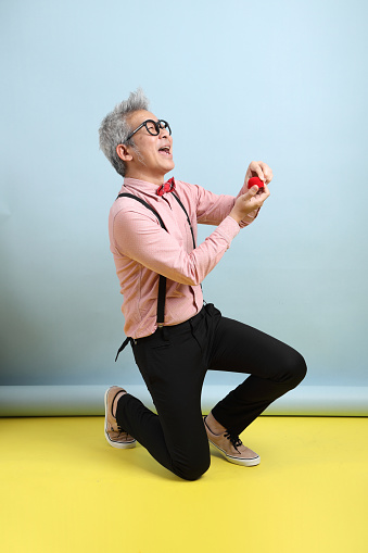 Asian senior man in black suspenders with red bow with gesture of  present an Engagement Ring isolated on blue and yellow background. St Valentine's Day