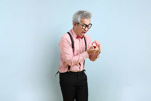 Asian senior man in black suspenders with red bow with gesture of  present an Engagement Ring isolated on blue and yellow background. St Valentine's Day