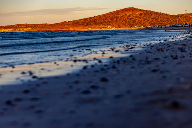 Coastal sunset with sea and sunlit mountain hills in background, stones in beach sand from lower perspective in blurred foreground, afternoon in Baja California Sur, Mexico
