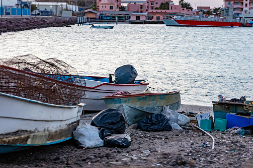 Environmental pollution on coastal beach, boats in sand, accumulation of trash, plastic bags, nets and ropes, metal fishing traps, buildings and boats in background, La Paz, Baja California Sur Mexico