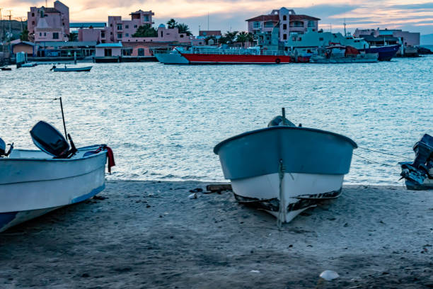 Coastal beach with front view of motorboat on sand, bay sea, buildings and boats in background, cloudy afternoon in La Paz, Baja California Sur Mexico