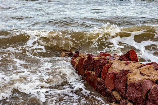 Small reddish brown irregular stone wall on beach, sea waves breaking on rocky coast, forming abundant foam, uneven surface, cloudy day