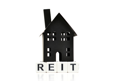 Closeup of a generic house figurine on top of letter blocks reading REIT, symbolizing a real estate investment trust.