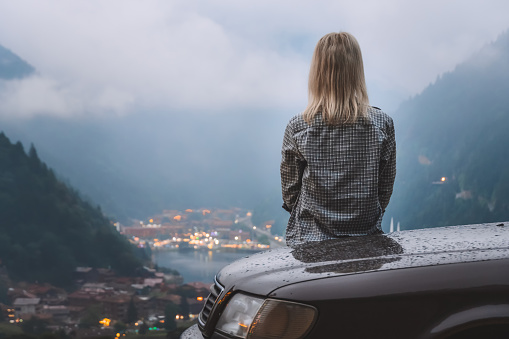 Young blonde woman in a plaid shirt is sitting in a car, admiring the beautiful view of the mountains and the lake while traveling on a cloudy day, focus is on the girl. Uzungol, Turkey.