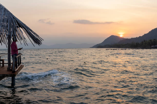 Mature man relaxes on pier above sea as sun rises over distant mountains, Phu Quoc island