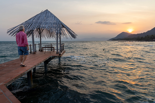 Mature man relaxes on pier above sea as sun rises over distant mountains, Phu Quoc island