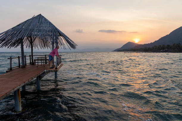 Mature man relaxes on pier above sea as sun rises over distant mountains, Phu Quoc island