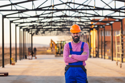 With a backdrop of machinery and construction materials, a male worker poses for a portrait, his attire reflecting the grit and determination required for his role on the site.