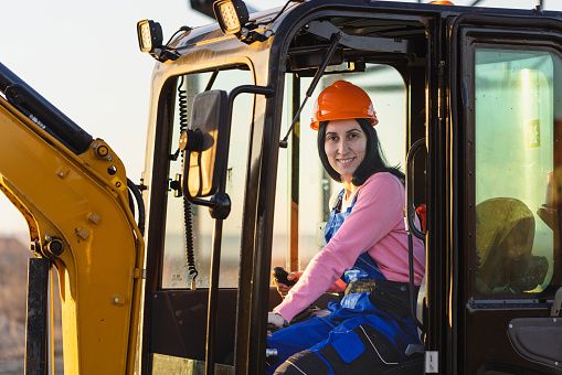 A female worker drives an excavator, skillfully maneuvering it to clear the construction area.