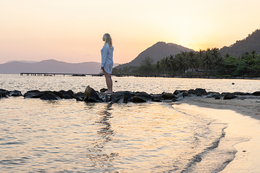 Mature woman steps between rocks on beach as sun rises over distant mountains