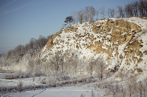 A stunning winter landscape of a snow-covered cliff with a lone tree, popular for hiking and rock climbing, offering breathtaking views of a frozen river and snowy forest.