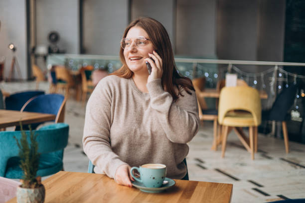 istock Attractive 30s female with cup of coffee having talking conversation with mobile phone while rest in cafe. Casual young woman using mobile phone indoors 1995636064