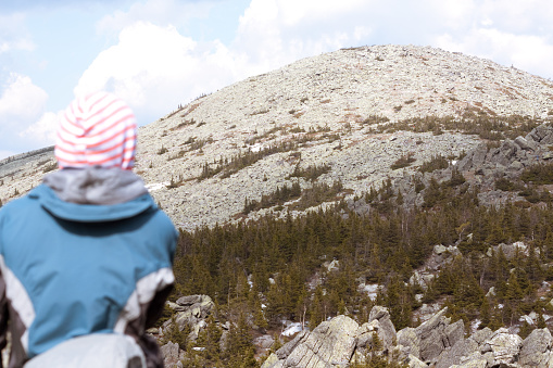 A person wearing a striped hat is looking at a rocky mountain peak. The mountain is covered in snow. A large number of rocks and boulders lie around the mountain.