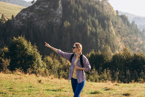Blonde 30s girl pointing finger on mountains background. Young female with backpack hiking in nature. Beautiful woman walking in sport clothes. Active lifestyle concept