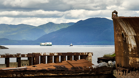 Old rusty ship hulls act as a breakwater.  Tug and barge coming down the inlet with distant mountains in the background.